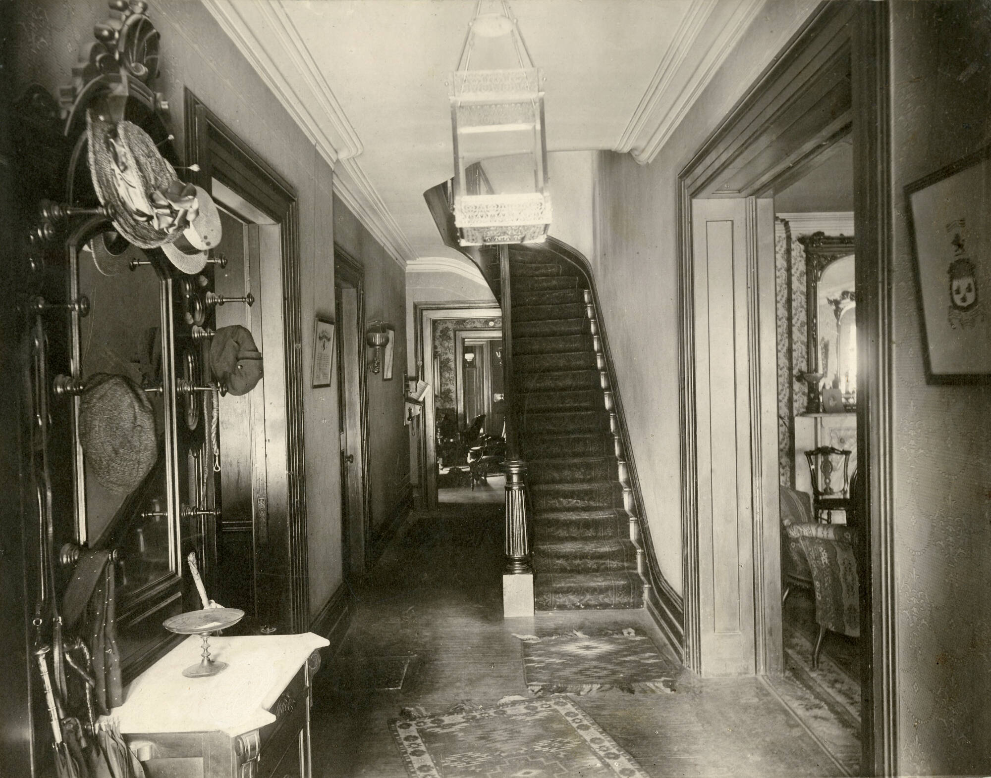 Photograph of the interior of a house. There is a staircase in the center of the image that leads upstairs, and there is a light fixture covering the view of the top of the stairs on the ceiling.